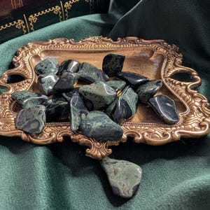 May include: A collection of polished green and black stones displayed on a decorative gold-colored tray. The tray sits on a dark green velvet surface, with a glimpse of antique books in the background. The stones vary in size and shape.
