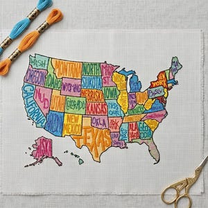 May include: A cross-stitch embroidery project on white fabric featuring a map of the United States with each state labeled in colorful thread. Two skeins of embroidery floss in blue and orange are positioned at the top left, and gold scissors are at the bottom right.