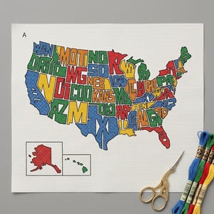 May include: A white needlepoint canvas featuring a map of the United States with states outlined in blue, green, red, and yellow, containing abbreviated state names. Includes a separate inset for Alaska and Hawaii, gold scissors, and embroidery floss.