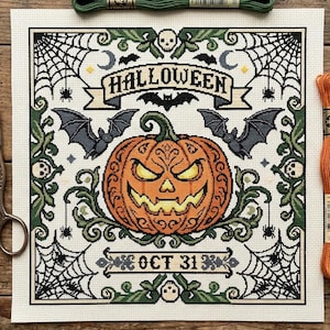 May include: A square cross-stitch pattern on white fabric featuring a central orange jack-o'-lantern, bats, spiderwebs, and the text HALLOWEEN and OCT 31. Four skeins of embroidery floss in blue, green, orange, and white, and metal scissors.