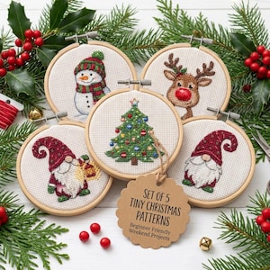 Op de afbeelding: Vijf ronde borduurringen met kruissteekontwerpen van een sneeuwpop, een rendier, een kerstboom en twee kabouters. Een bruin label vermeldt SET OF 5 TINY CHRISTMAS PATTERNS Beginner Friendly Projects. Omringd door hulst en dennentakken.