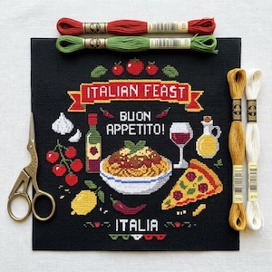 Pode incluir: Um tecido de ponto cruz preto com um design de banquete italiano com o texto ITALIAN FEAST, BUON APPETITO e ITALIA. O padrão inclui massa, pizza, vinho, azeite, tomates, alho e limão, acompanhado por tesouras de bordar e quatro meadas de fio.