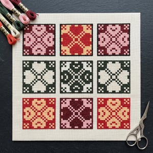 May include: A completed cross-stitch sampler featuring nine squares with floral patterns in various colors, including pink, red, green, and cream. Embroidery floss in coordinating colors and small scissors are visible. The sampler is on a neutral-colored fabric.