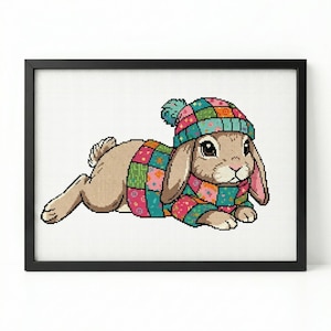Op de afbeelding: Een ingelijste print met een bruin konijn met een kleurrijke patchwork hoed en trui. Het konijn ligt, met zijn oren omhoog. Het kunstwerk heeft een kruissteek-uiterlijk, tegen een witte achtergrond.