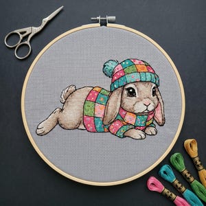 May include: A cross-stitch embroidery project in a round wooden hoop featuring a brown lop-eared rabbit wearing a colorful patchwork sweater and matching beanie with a pom-pom. The design is stitched on gray fabric, accompanied by metal scissors.