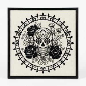 May include: A framed cross-stitch featuring a Day of the Dead skull design in black and white. The skull is encircled by black roses, flowers, and a decorative border. The background is a light beige fabric.