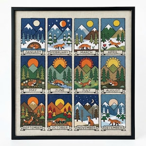 May include: A framed cross-stitch depicting the months of the year, each with a fox in a seasonal landscape. The artwork features mountains, trees, and varying weather conditions, with text labels for each month.
