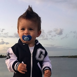 Peut inclure: Un tout-petit portant une veste de baseball bleu et blanche avec un grand 'C' sur le côté droit, un jean bleu et des baskets noires. Le tout-petit a une coupe de cheveux en mohawk et porte une sucette bleue.