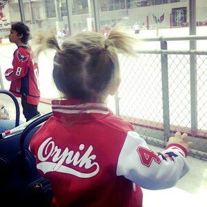 Pu&ograve; includere: Un bambino piccolo indossa una giacca da baseball rossa e bianca con il nome "Orpik" sul retro. Il bambino sta guardando una partita di hockey su una pista di ghiaccio.