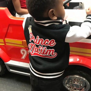 May include: A black and white baseball jacket with a red and white embroidered design that says "Prince Na'iim". The jacket has a white collar and white sleeves. The jacket is being worn by a child.