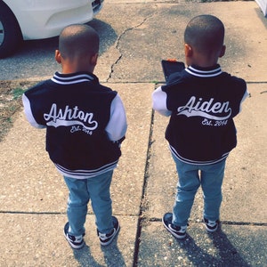 Pode incluir: Dois meninos pequenos vestindo jaquetas de beisebol pretas e brancas com seus nomes, Ashton e Aiden, e o ano de 2014 nas costas. Eles est&atilde;o usando jeans azul e t&ecirc;nis pretos.