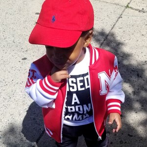Może przedstawiać: Małe dziecko w czerwono-białej kurtce bejsbolowej z białym t-shirtem z napisem "Used A Ton Mama" pod spodem. Dziecko ma r&oacute;wnież czerwoną czapkę bejsbolową z niebieskim logo, czarne spodnie i czarno-białe buty sportowe.