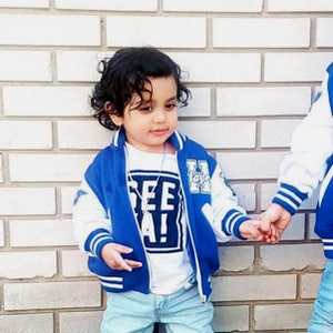 Może przedstawiać: Dw&oacute;ch młodych chłopc&oacute;w w niebieskich i białych kurtkach bejsbolowych. Chłopiec po lewej stronie ma na sobie białą koszulkę z nadrukiem "See Ya!" Chłopiec po prawej stronie ma na sobie białą koszulkę z nadrukiem "Styli".