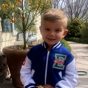 Pu&ograve; includere: Un bambino piccolo che indossa una giacca da baseball blu e bianca con la scritta "Esopi" sul davanti. Sta tenendo un guanto da baseball in pelle marrone con un logo "Wilson" bianco.