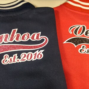 Może przedstawiać: Dwie bejsbol&oacute;wki w kolorze granatowym i czerwonym z białymi paskami na kołnierzu. Kurtki mają biały i błyszczący wz&oacute;r z tyłu z nazwami "Ainhoa" i "Vega" oraz tekstem "Est. 2016".