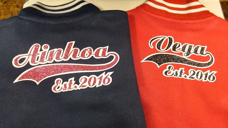 Może przedstawiać: Dwie bejsbol&oacute;wki w kolorze granatowym i czerwonym z białymi paskami na kołnierzu. Kurtki mają biały i błyszczący wz&oacute;r z tyłu z nazwami "Ainhoa" i "Vega" oraz tekstem "Est. 2016".