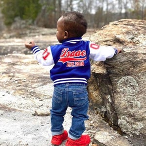 May include: A toddler wearing a blue and white baseball jacket with red lettering that says "Isaac EST. 2018". The jacket has white sleeves with blue and white stripes at the cuffs. The toddler is wearing blue jeans and red sneakers.