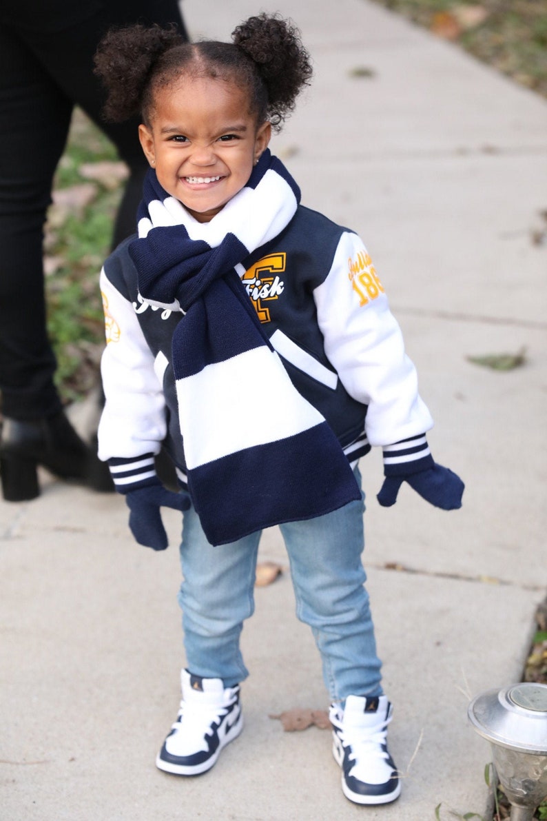 Może przedstawiać: Mała dziewczynka w niebiesko-białej szaliku, granatowej i białej kurtce bejsbolowej z napisem "Frisk" i "186", niebieskich dżinsach i białych butach sportowych z granatowymi akcentami.