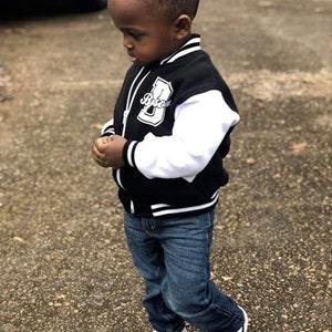 Pode incluir: Um menino pequeno vestindo uma jaqueta de beisebol preta e branca com a letra "B" na frente. Ele tamb&eacute;m est&aacute; vestindo jeans azul e t&ecirc;nis preto e branco.