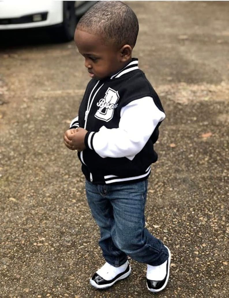 Pode incluir: Um menino pequeno vestindo uma jaqueta de beisebol preta e branca com a letra "B" na frente. Ele tamb&eacute;m est&aacute; vestindo jeans azul e t&ecirc;nis preto e branco.