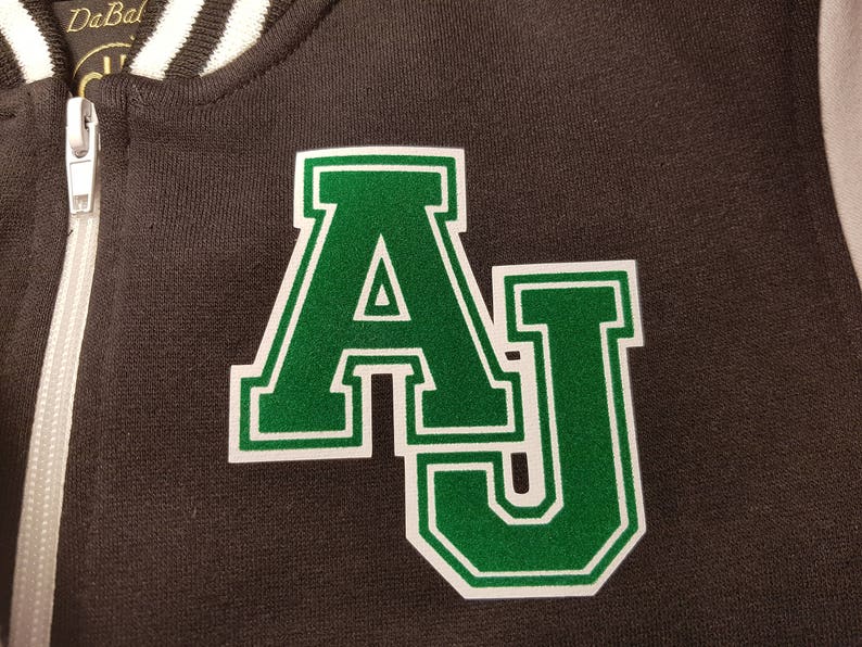 Może przedstawiać: Czarna kurtka bomberowa z białym zamkiem błyskawicznym i białym wykończeniem wok&oacute;ł kołnierza. Kurtka ma zieloną naszywkę z filcu z literami "AJ" w kolorze białym.