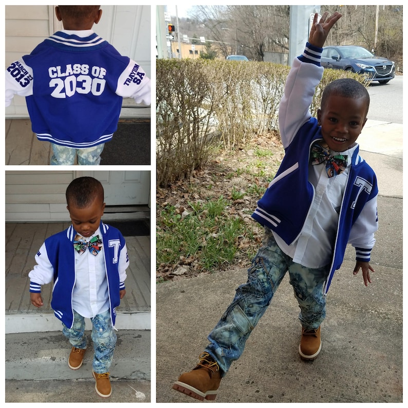May include: A young boy wearing a blue and white varsity jacket with the text "Class of 2030" on the back. He is also wearing a white shirt, blue jeans, and a colorful bow tie.