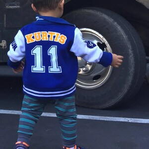 May include: A young boy wearing a blue and white baseball jacket with the name "Kurtis" and the number "11" on the back. He is standing in front of a large vehicle.