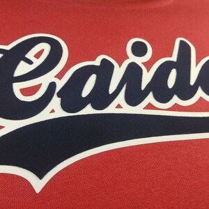 Może przedstawiać: Czerwona bluza z białym obramowaniem i granatowym napisem "Caide" w ozdobnym, pisanym stylu.