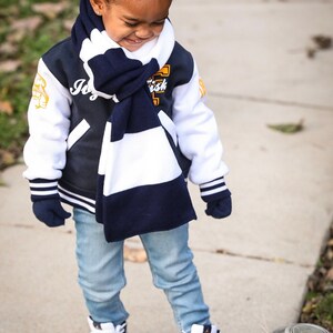 Może przedstawiać: Młoda osoba w niebiesko-białej szaliku, granatowej i białej kurtce varsity z napisem "Joy" z przodu, niebieskich dżinsach i biało-granatowych butach sportowych.