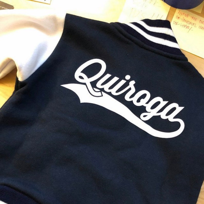 Pode incluir: Uma jaqueta de beisebol azul marinho e branca com o nome "Quiroga" em letras brancas nas costas.