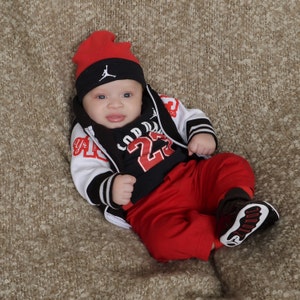May include: A baby boy wearing a black and white baseball jacket with red accents, a black and red shirt with the number 23, and red pants. He is wearing a red and black baseball cap with a white logo.