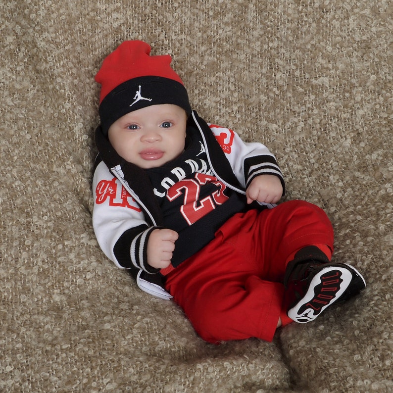 May include: A baby boy wearing a black and white baseball jacket with red accents, a black and red shirt with the number 23, and red pants. He is wearing a red and black baseball cap with a white logo.