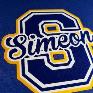 Pode incluir: Um remendo de feltro azul e amarelo com um contorno branco do n&uacute;mero 6 e a palavra "Simeon" em letras brancas cursivas.