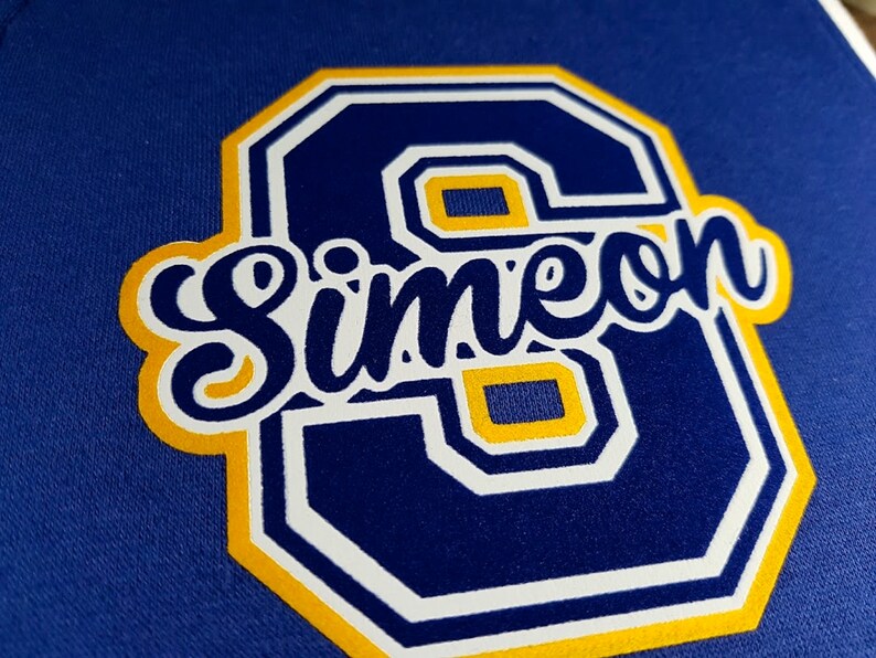 Pode incluir: Um remendo de feltro azul e amarelo com um contorno branco do n&uacute;mero 6 e a palavra "Simeon" em letras brancas cursivas.