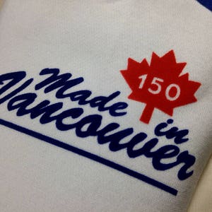 May include: White fabric with the text "Made in Vancouver" in blue script, a red maple leaf with "150" inside, and blue trim. The design celebrates Vancouver's 150th anniversary.
