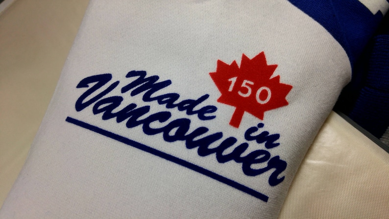 May include: White fabric with the text "Made in Vancouver" in blue script, a red maple leaf with "150" inside, and blue trim. The design celebrates Vancouver's 150th anniversary.
