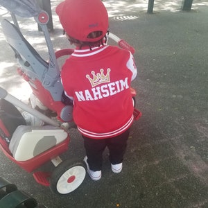 May include: A child wearing a red and white baseball jacket with a gold crown and the name "NAHSEIM" embroidered on the back. The child is wearing a red baseball cap with the word "POLO" on it and is standing next to a red and white tricycle.