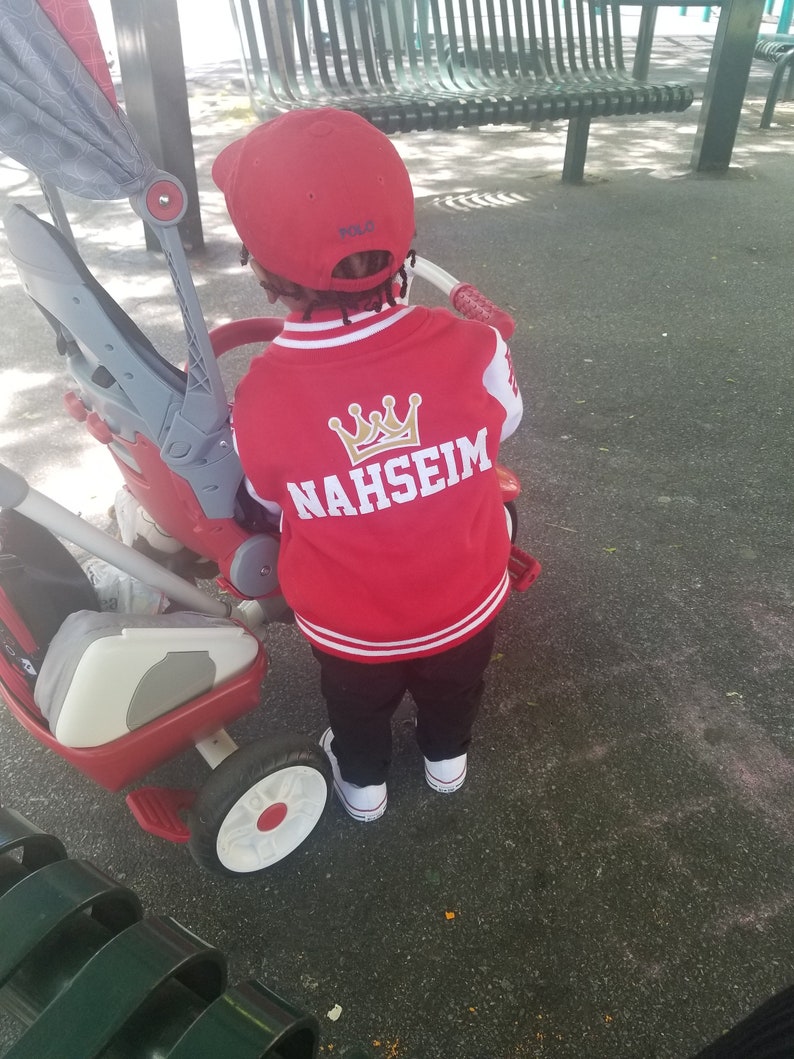 May include: A child wearing a red and white baseball jacket with a gold crown and the name "NAHSEIM" embroidered on the back. The child is wearing a red baseball cap with the word "POLO" on it and is standing next to a red and white tricycle.