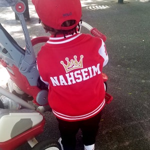 Może przedstawiać: Czerwono-biała kurtka bejsbolowa z koroną w kolorze złotym i haftowanym napisem "NAHSEIM" na plecach. Kurtkę nosi dziecko w czerwonej czapce bejsbolowej z napisem "POLO".
