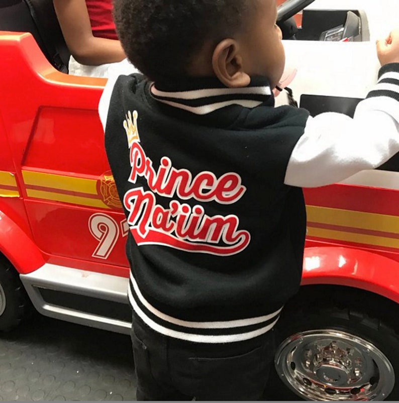 May include: A black and white baseball jacket with a red and white embroidered design that says "Prince Na'iim". The jacket has a white collar and white sleeves. The jacket is being worn by a child.