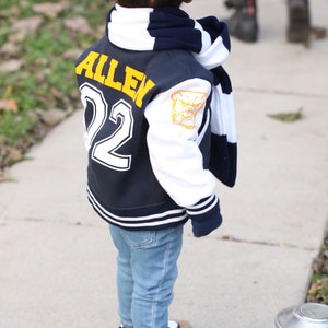 Może przedstawiać: Młoda osoba w niebiesko-białej kurtce bejsbolowej z imieniem "Allen" i numerem "22" na plecach. Kurtka ma biały pasek na rękawach i ż&oacute;łtego buldoga na prawym rękawie. Osoba nosi r&oacute;wnież niebieskie dżinsy i granatowo-białe buty sportowe.