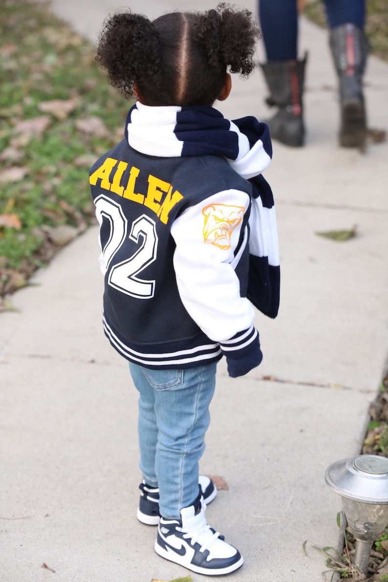 Może przedstawiać: Młoda osoba w niebiesko-białej kurtce bejsbolowej z imieniem "Allen" i numerem "22" na plecach. Kurtka ma biały pasek na rękawach i ż&oacute;łtego buldoga na prawym rękawie. Osoba nosi r&oacute;wnież niebieskie dżinsy i granatowo-białe buty sportowe.