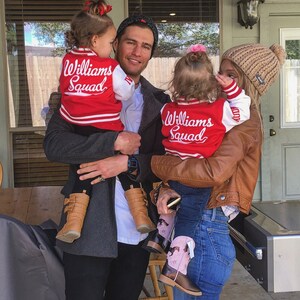 May include: A family poses outdoors, featuring two young children wearing red and white letterman jackets with "Williams Squad" on the back. The jackets are paired with black leggings and brown boots. The adults are wearing casual attire.