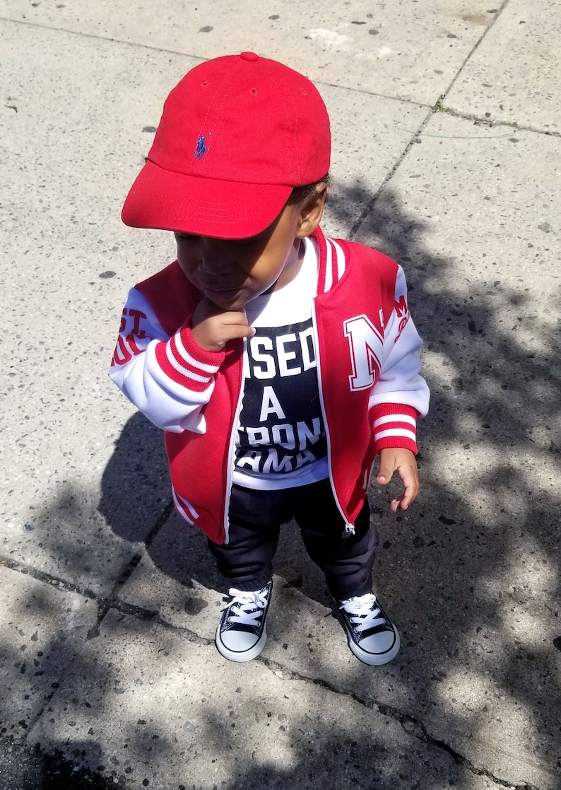 Może przedstawiać: Małe dziecko w czerwono-białej kurtce bejsbolowej z białym t-shirtem z napisem "Used A Ton Mama" pod spodem. Dziecko ma r&oacute;wnież czerwoną czapkę bejsbolową z niebieskim logo, czarne spodnie i czarno-białe buty sportowe.