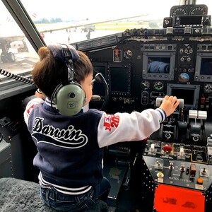 Może przedstawiać: Małe dziecko w niebiesko-białej kurtce bejsbolowej z napisem "Darwin" na plecach siedzi w kokpicie samolotu. Dziecko ma na sobie słuchawki i trzyma rękę na dźwigni sterującej. Kokpit jest wypełniony r&oacute;żnymi wskaźnikami i elementami sterującymi.