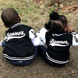 Pode incluir: Duas crian&ccedil;as vestindo jaquetas de beisebol pretas e brancas com o nome "Bonner" e "Est. 2021" nas costas. As jaquetas t&ecirc;m mangas brancas e corpos pretos com listras brancas ao redor da parte inferior.