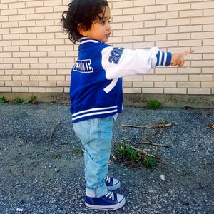 May include: A young child wearing a blue and white baseball jacket with the text "Rookie" on the back and "2014" on the sleeve. The child is wearing blue jeans and blue sneakers.