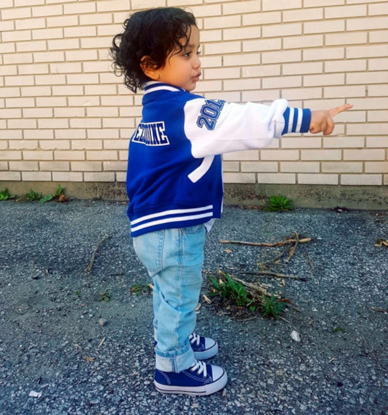 May include: A young child wearing a blue and white baseball jacket with the text "Rookie" on the back and "2014" on the sleeve. The child is wearing blue jeans and blue sneakers.