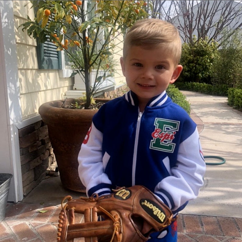 Pu&ograve; includere: Un bambino piccolo che indossa una giacca da baseball blu e bianca con la scritta "Esopi" sul davanti. Sta tenendo un guanto da baseball in pelle marrone con un logo "Wilson" bianco.