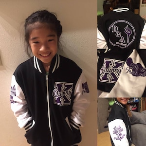 May include: A black and white varsity jacket with purple lettering. The jacket has a zipper closure and two pockets. The left sleeve has the text "Girls Competitive Team" and a flower design. The right sleeve has the text "Quinn K" and the text "Mary Trained". The back of the jacket has a circle with the text "DGA" and a silhouette of a gymnast.